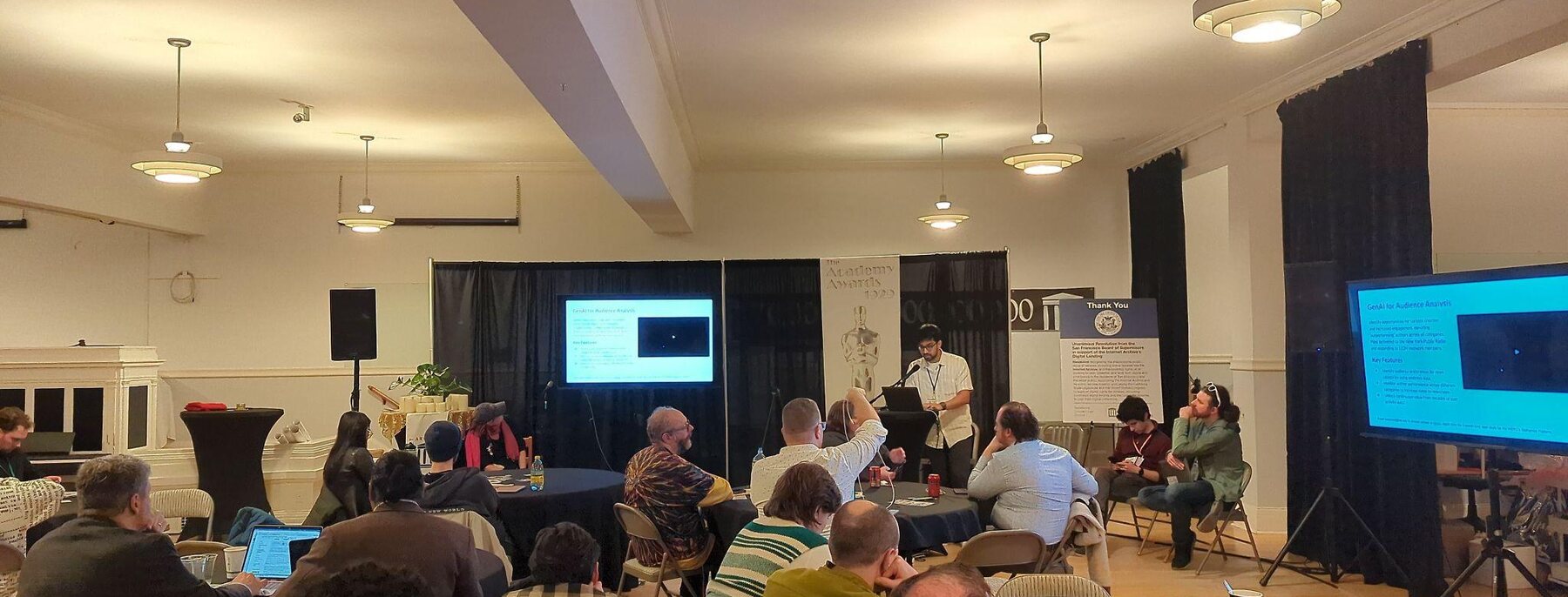 A room of participants seated at tables during a working session, with presentation screens at the front